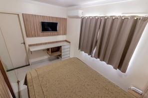 Quarto com cama, televisão montada em painel de madeira, janela com cortinas e ar condicionado embutido.