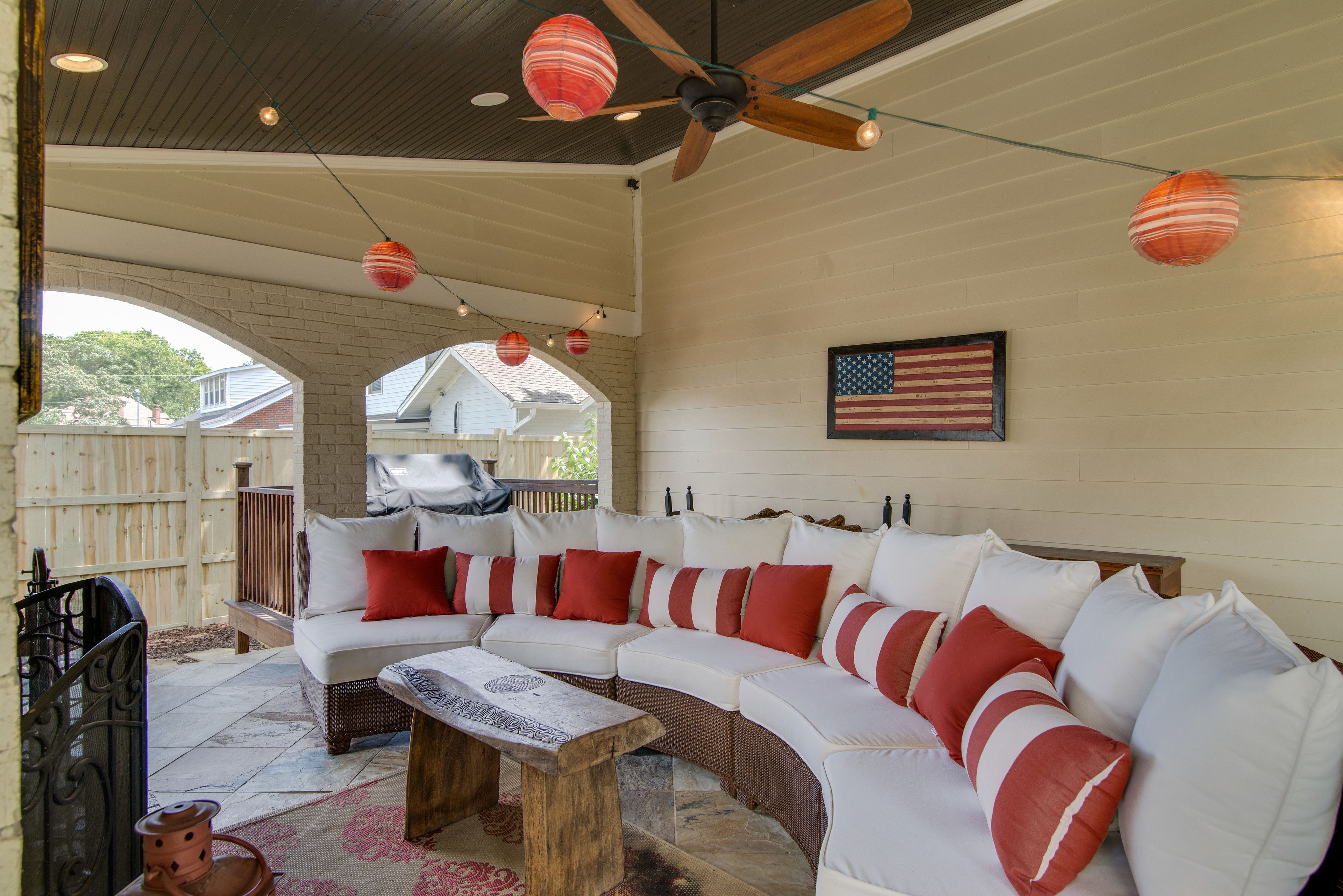 Un patio couvert comprenant un ensemble de meubles de jardin avec un canapé blanc incurvé, des coussins rouges et blancs, une table basse en bois et un ventilateur de plafond.