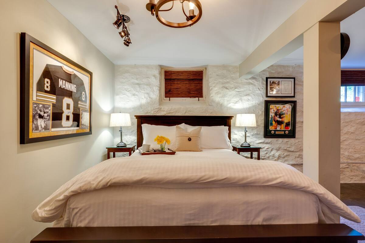 A neatly made bed with a large framed sports jersey, a wooden headboard, and a small window with blinds.