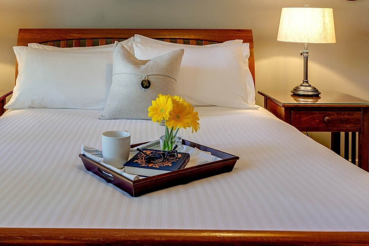 A neatly made bed with white linens, a tray with a mug, glasses, and flowers, and a bedside lamp.