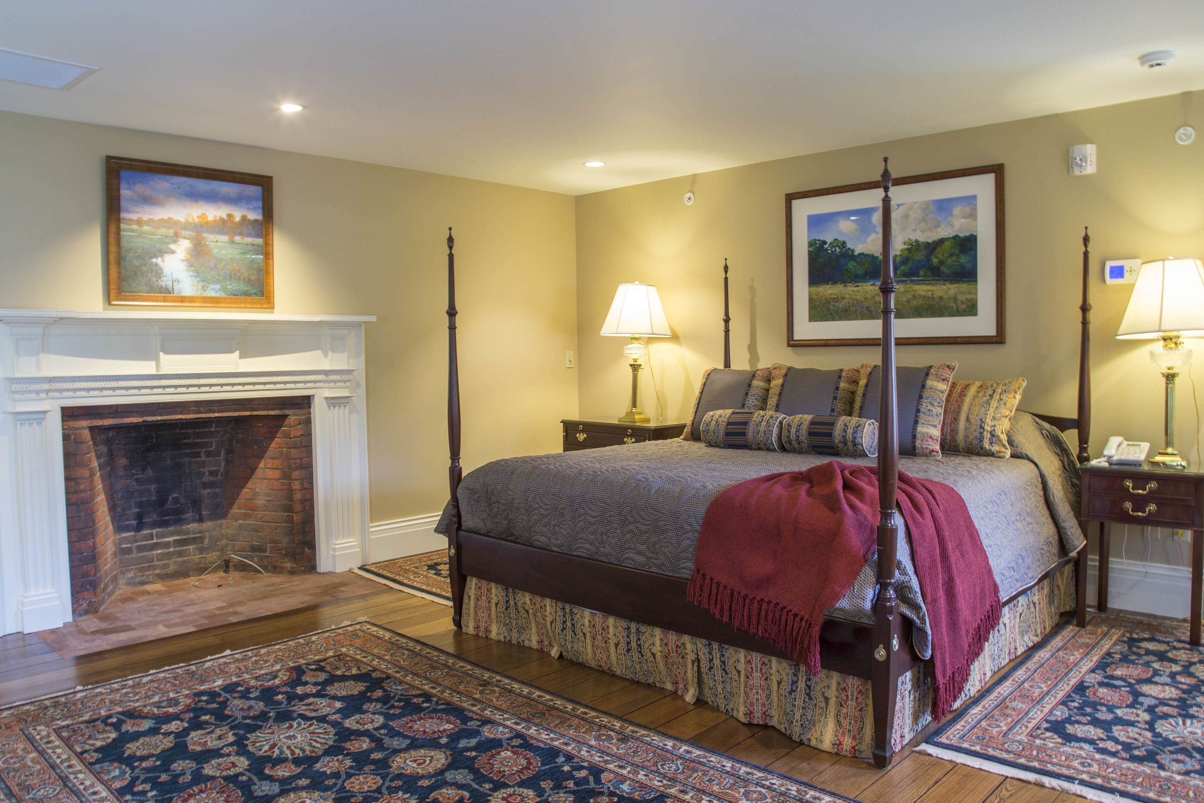 A four-poster bed with a red throw, a fireplace, two bedside lamps, and a large landscape painting above the bed.