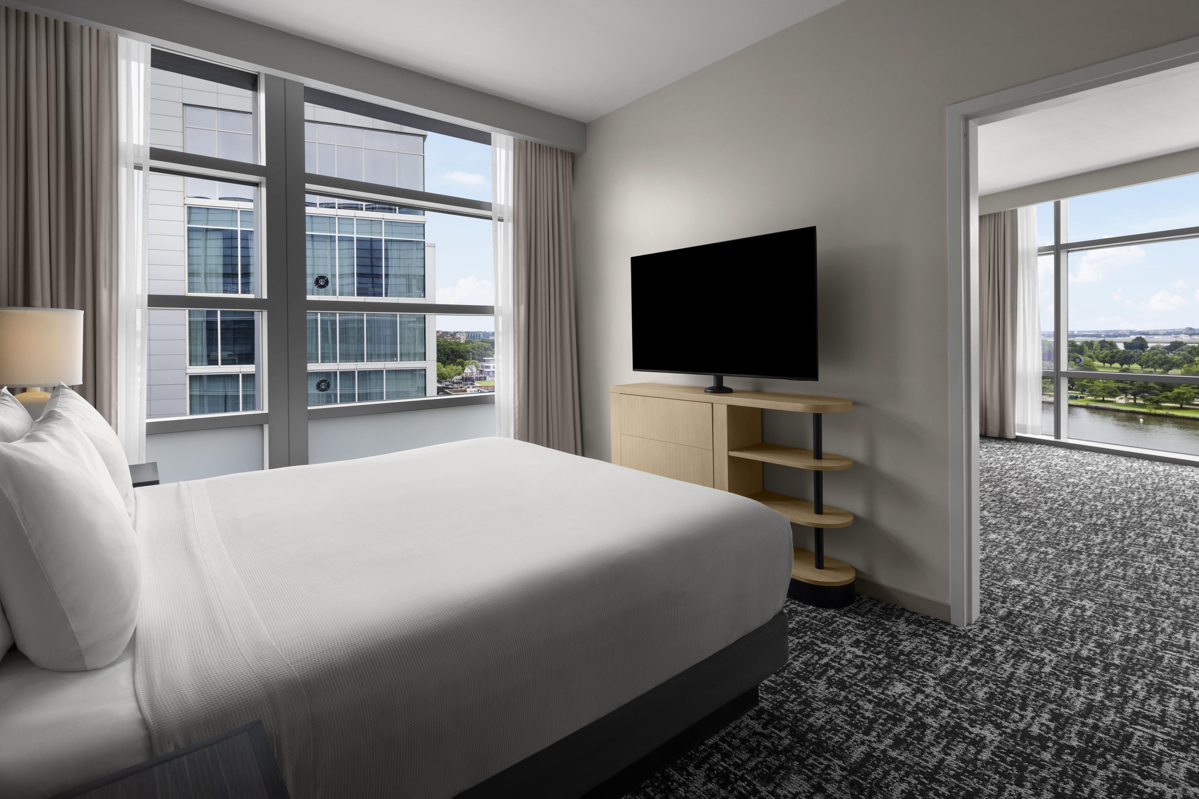 A hotel room with a bed, a TV on a wooden stand, a window with a view of buildings, and a carpeted floor.