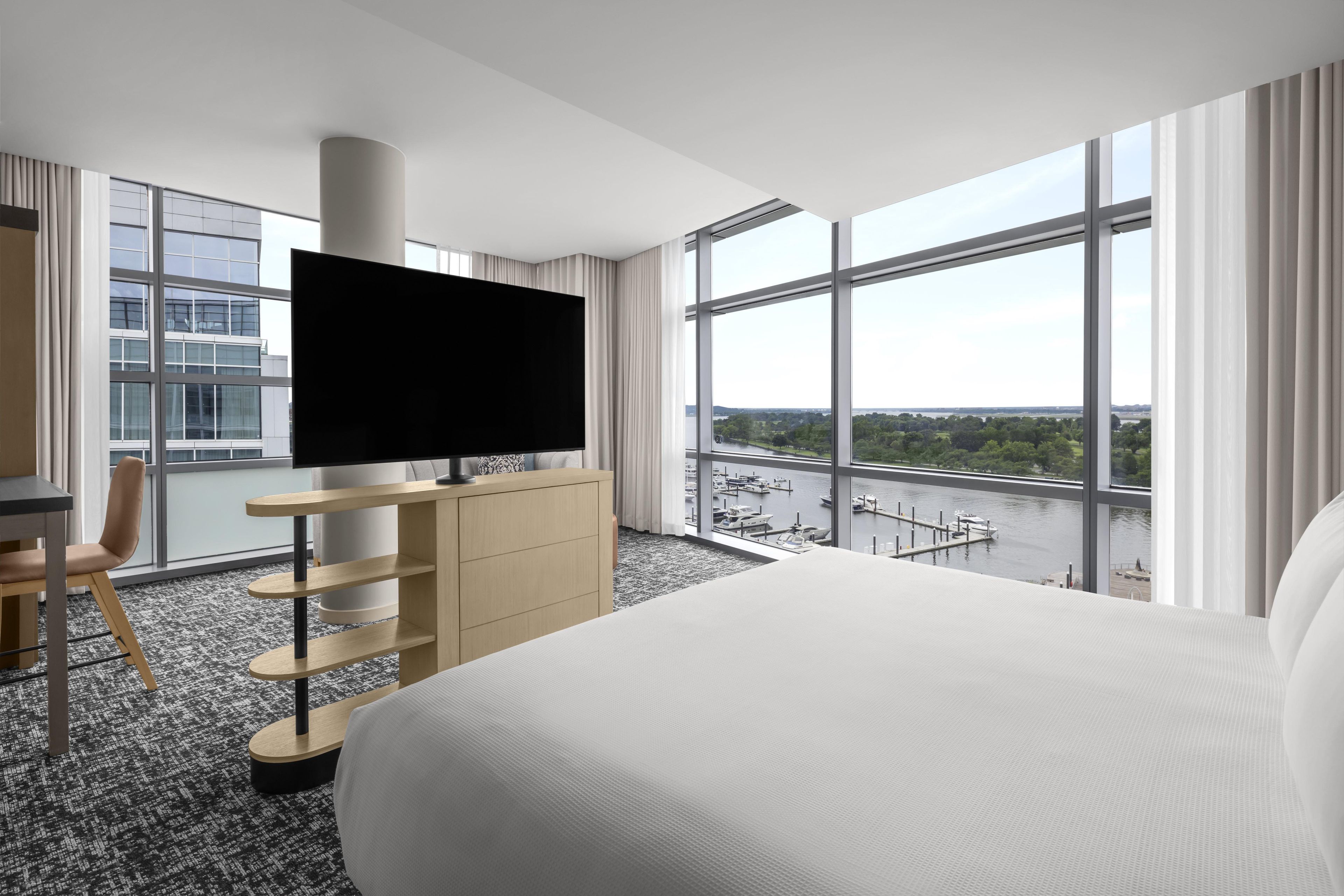 A hotel room with a large bed, a TV on a stand, a desk, and a view of a marina through large windows.