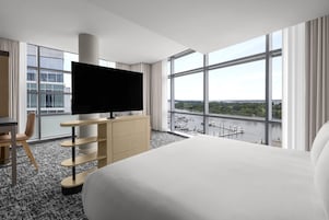 A hotel room with a large bed, a TV on a stand, a desk, and a view of a marina through large windows.