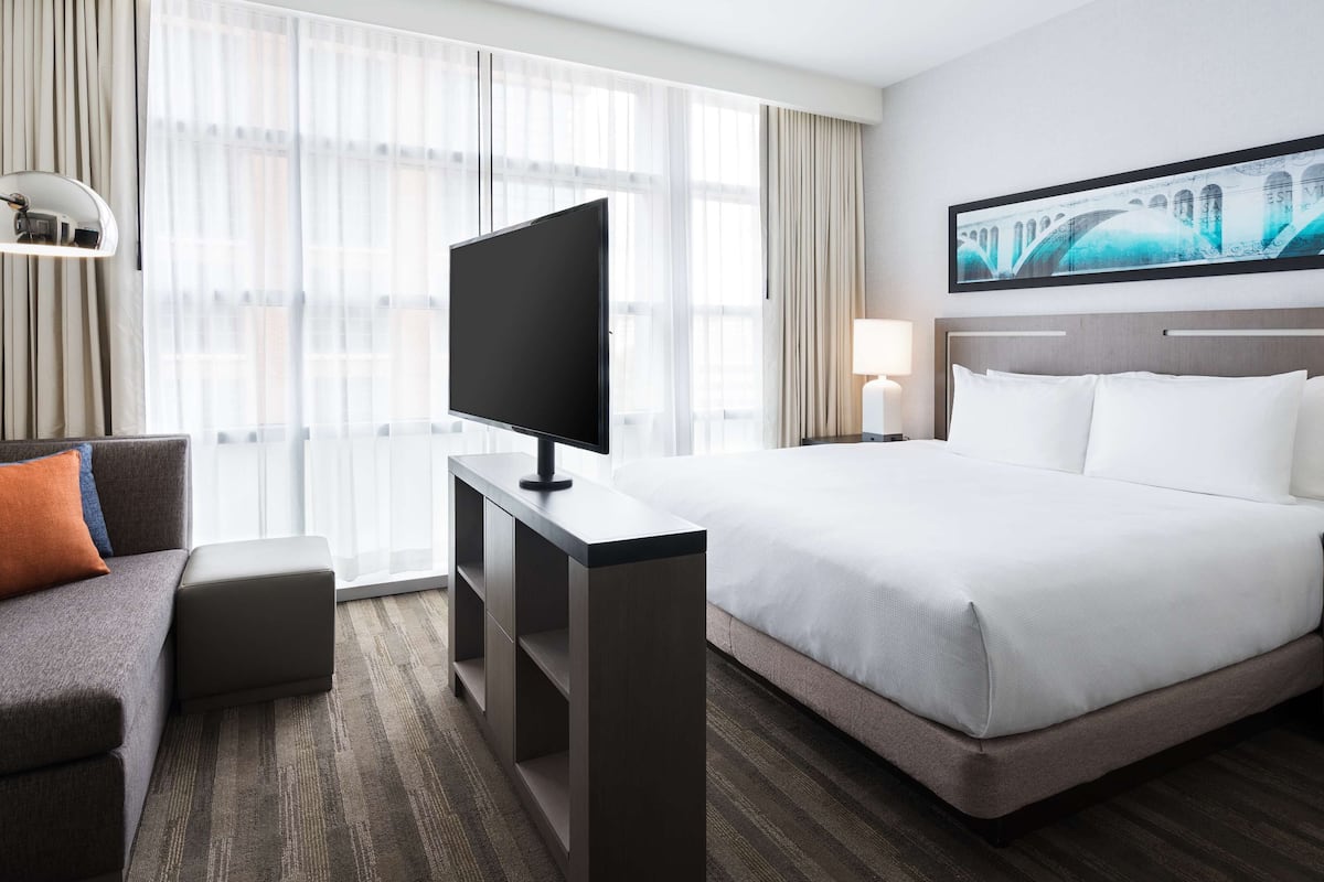 Habitación de hotel con una cama grande, un escritorio con televisor de pantalla plana, un sofá con cojines y un cuadro en la pared.