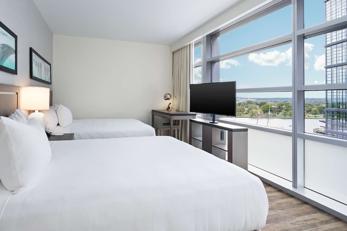 Habitación de hotel con dos camas, un escritorio con televisor y ventanas amplias con vistas a la ciudad.