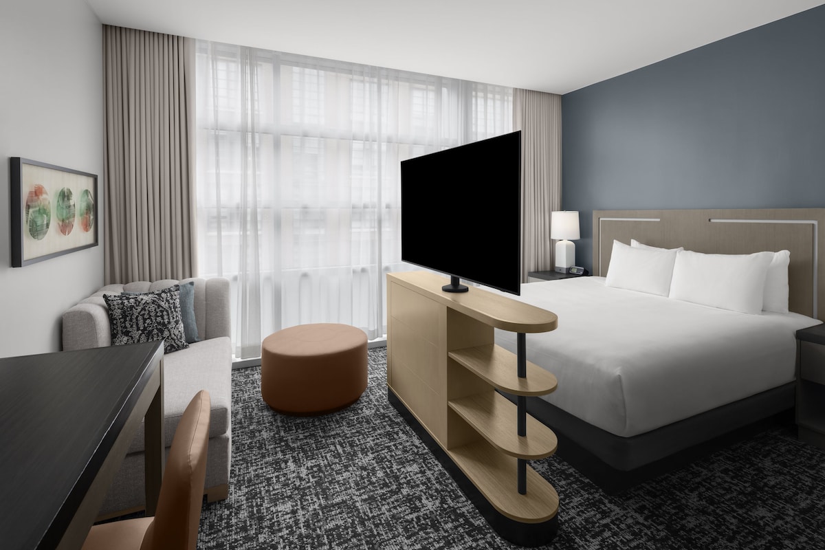 A hotel room with a bed, a TV mounted on a wooden console, a desk, a chair, and a large window with curtains.