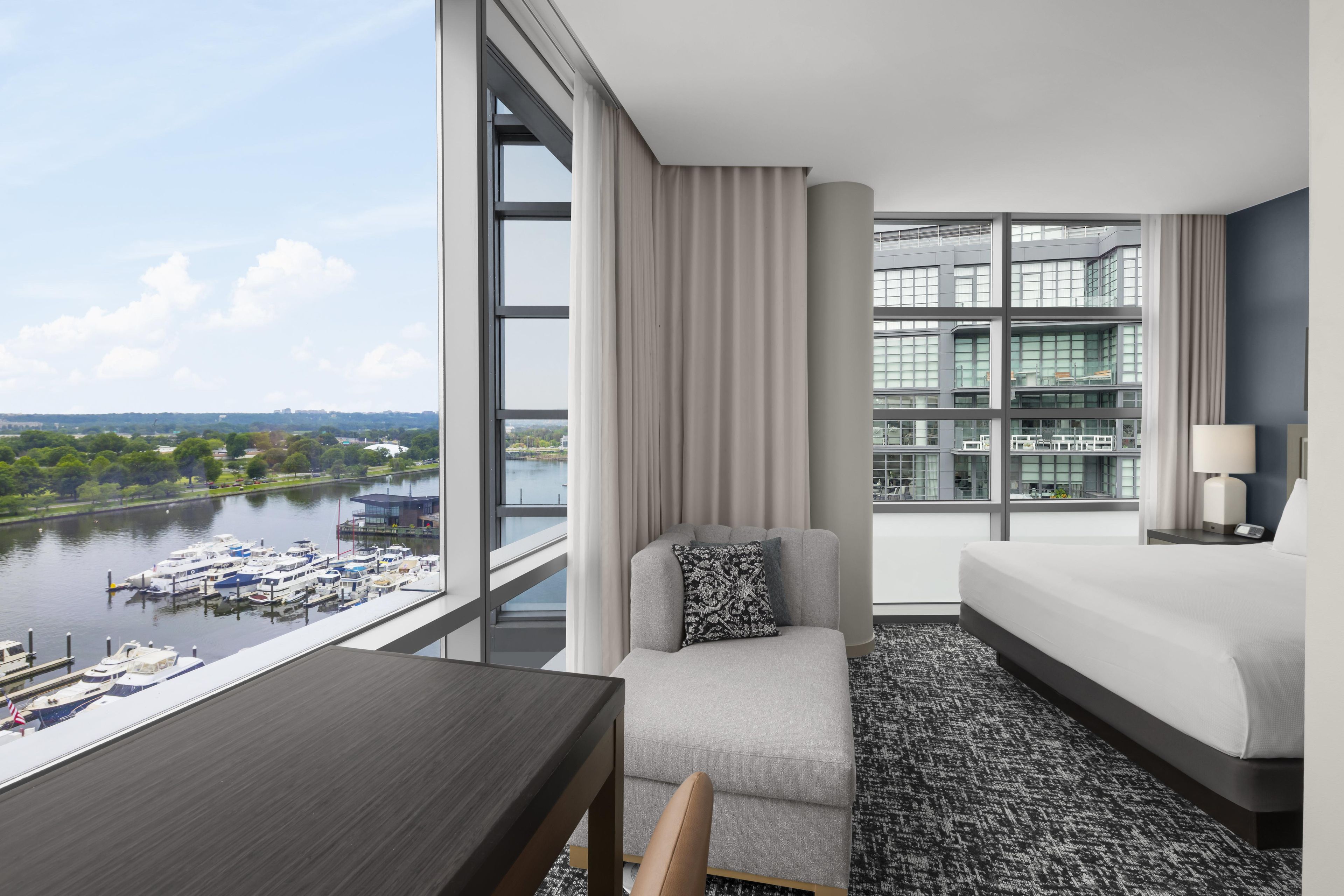 A modern hotel room with a large window, a sofa, a table, and a view of a river and boats.