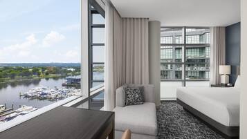 A modern hotel room with a large window, a sofa, a table, and a view of a river and boats.