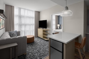 Habitación de hotel moderna con sofá, un taburete redondo pequeño, televisor de pantalla plana, escritorio con silla y una lámpara colgante.