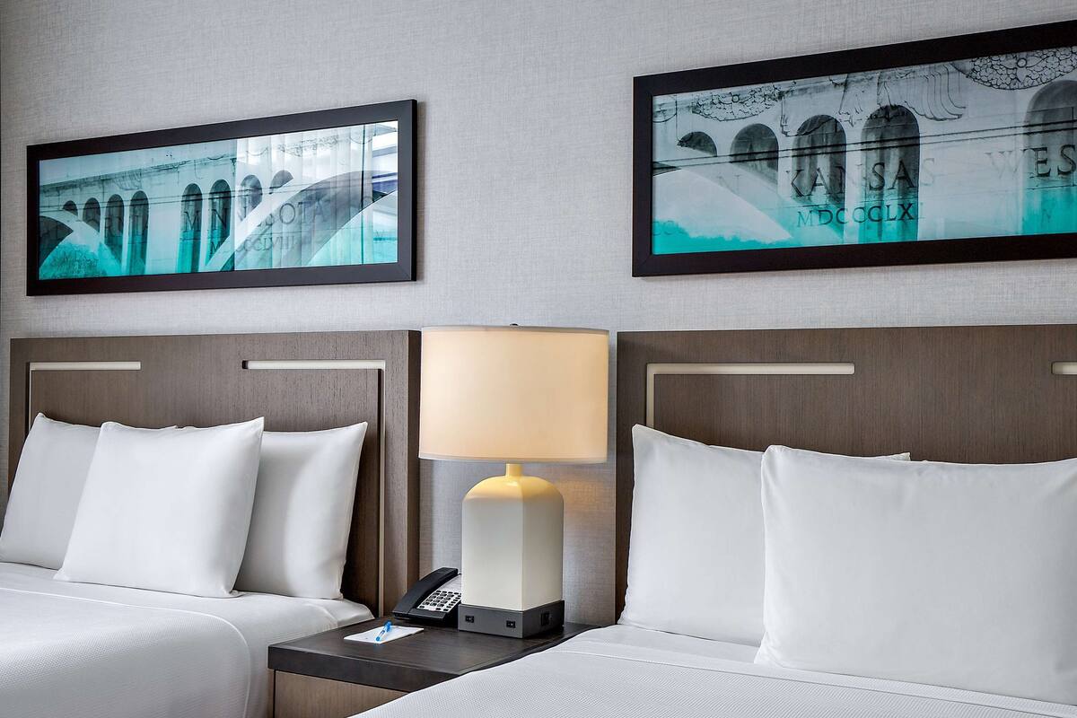Habitación de hotel con dos camas, una mesita de noche con lámpara y dos cuadros en la pared.