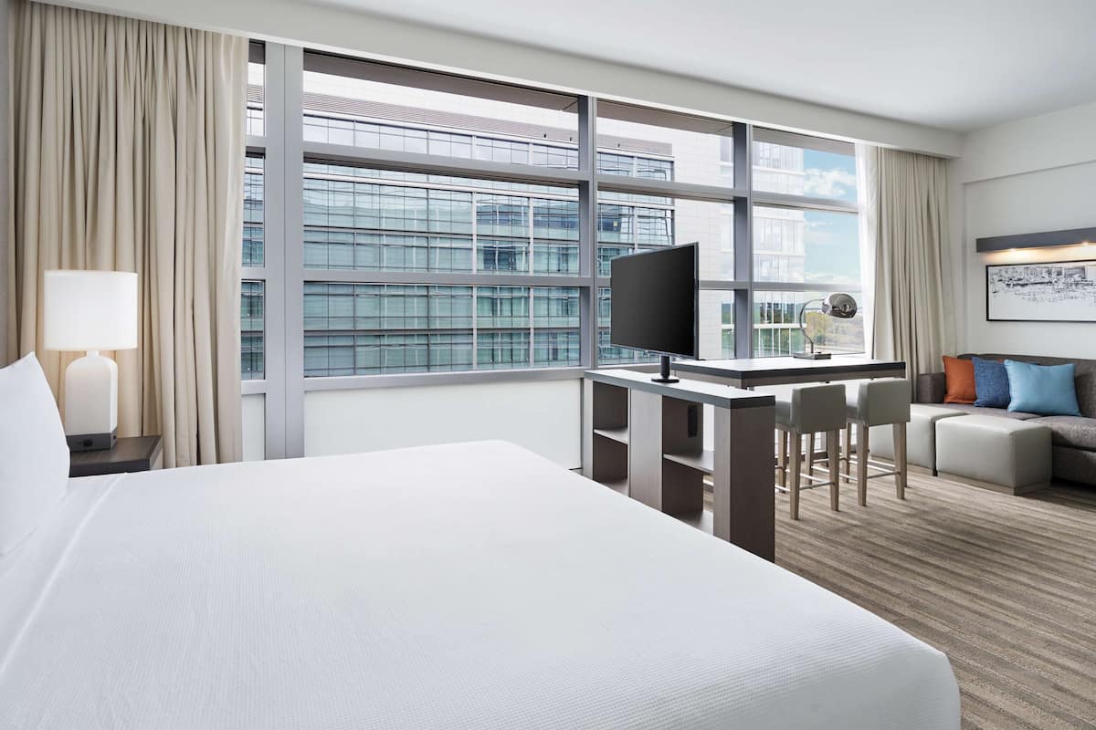 Habitación de hotel con una cama grande, un escritorio con silla, un sofá y un ventanal con vista a la ciudad.