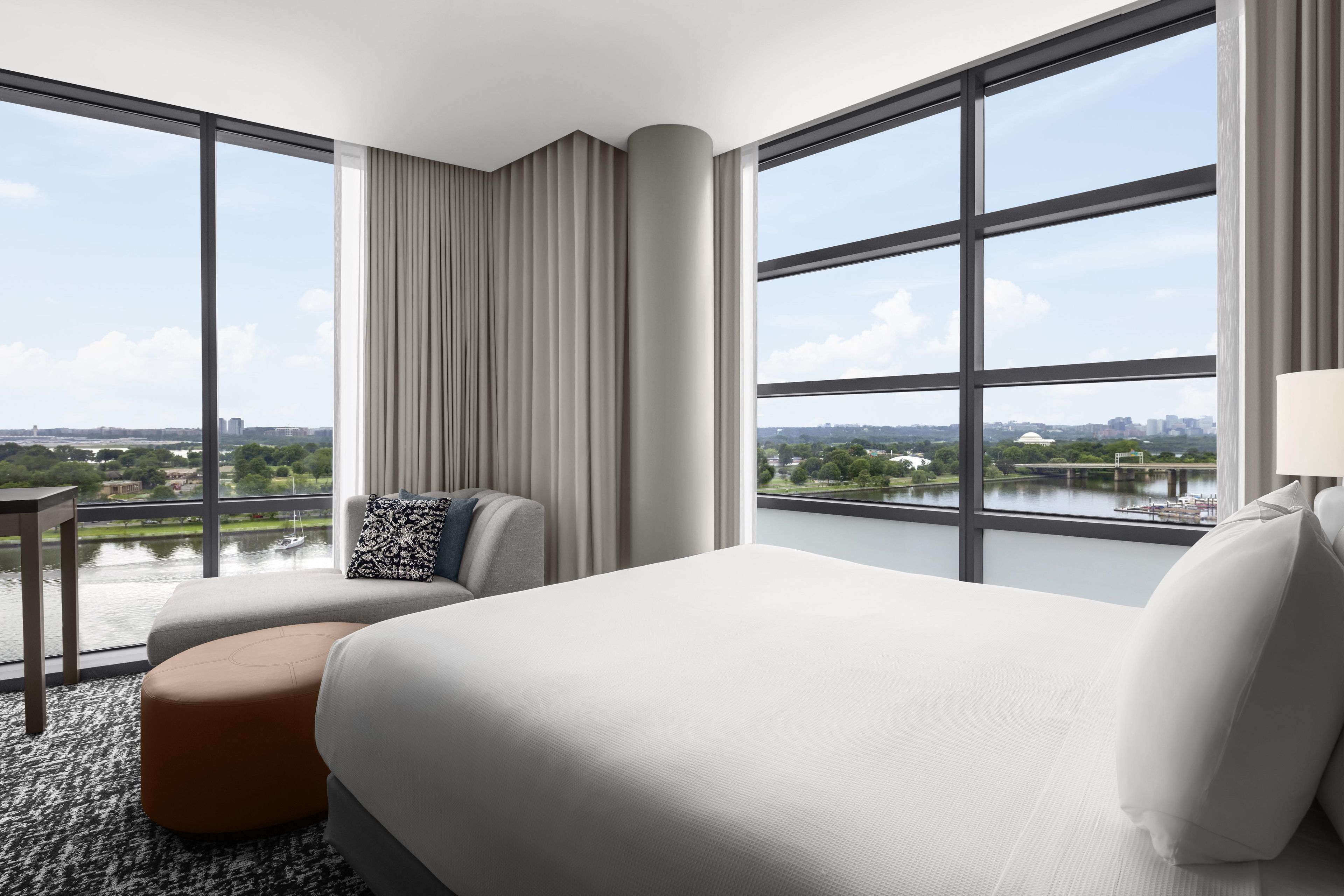 Ein Hotelzimmer mit einem großen Bett, einer Sitzecke inklusive Sessel und Hocker, einem kleinen Tisch und großen Fenstern mit Blick auf eine Stadtlandschaft und ein Gewässer.