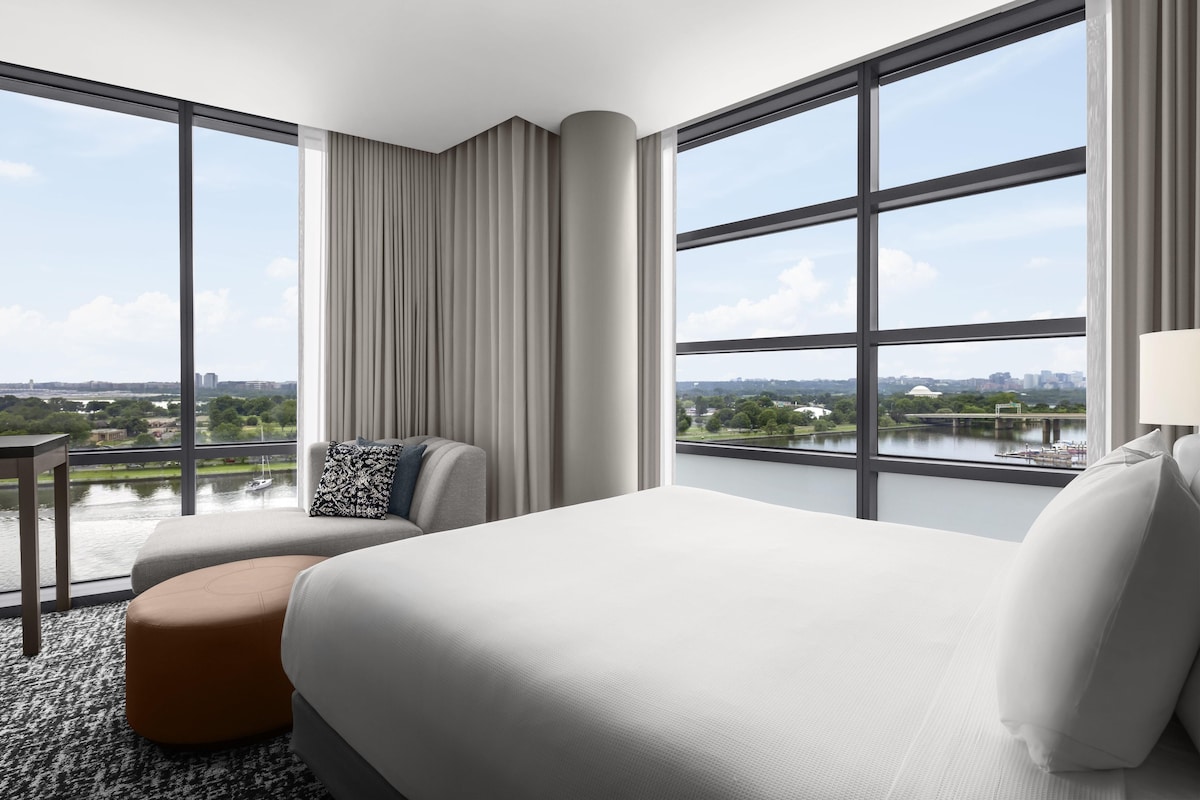 A hotel room with a large bed, a seating area with a chair and ottoman, a small table, and large windows offering a view of a cityscape and a body of water.