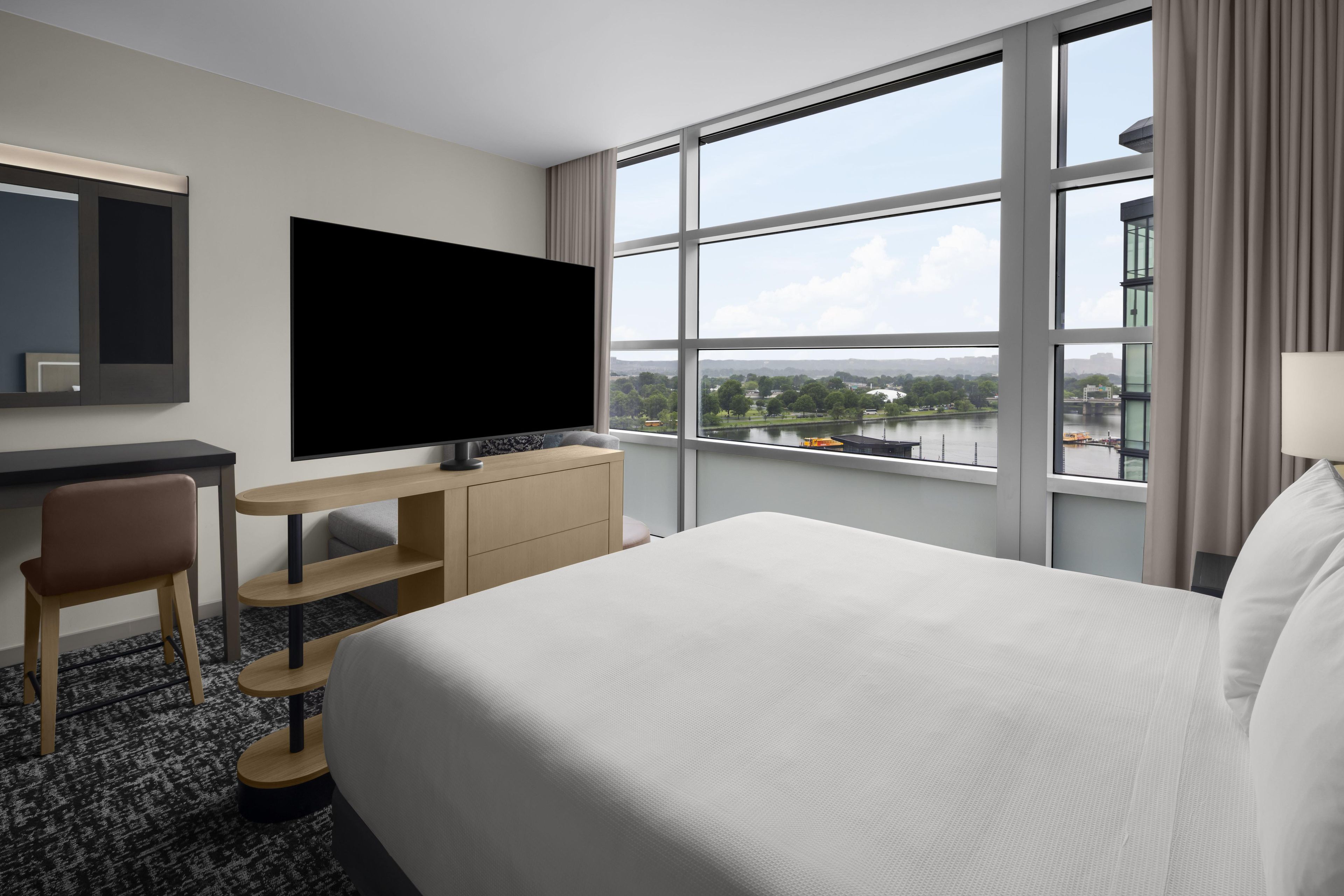 A hotel room with a bed, a desk with a flat-screen TV, a chair, and a large window offering a view of a river and cityscape.