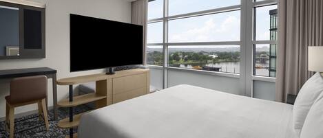 A hotel room with a bed, a desk with a flat-screen TV, a chair, and a large window offering a view of a river and cityscape.