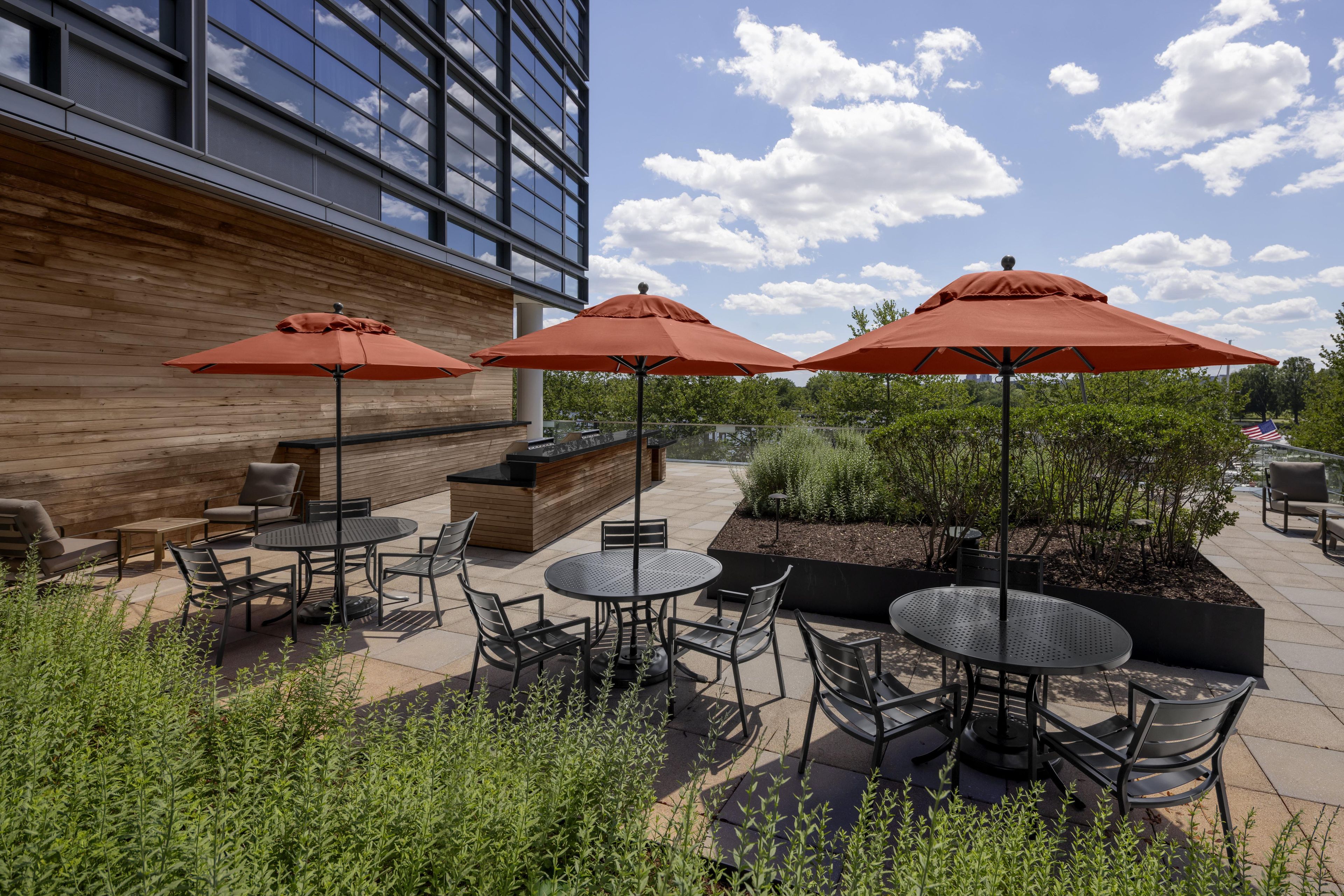An outdoor patio area with multiple tables and chairs, surrounded by greenery and a wooden wall.