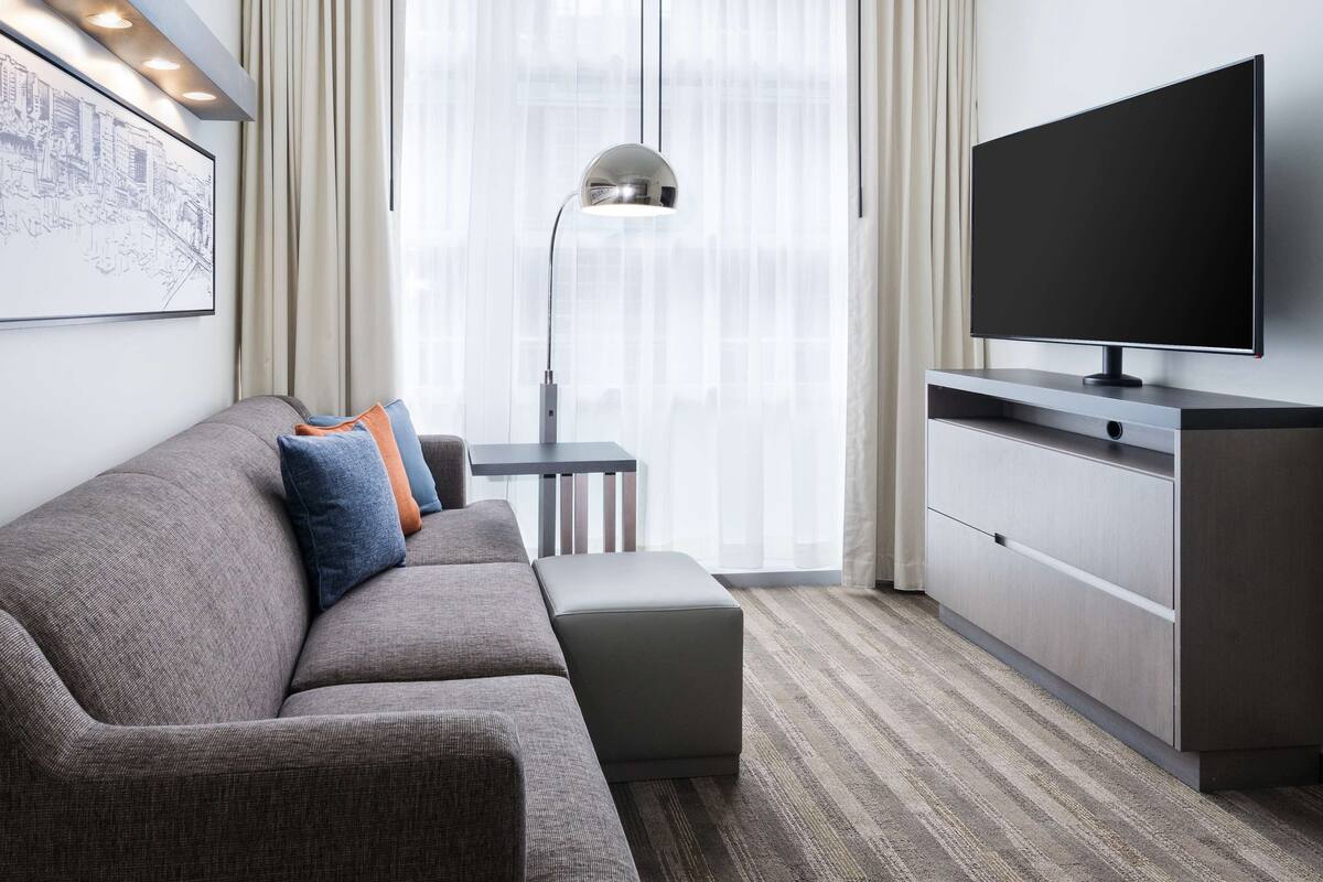 Una habitación de hotel moderna con un sofá gris, una mesita auxiliar pequeña, un televisor de pantalla plana y un ventanal con cortinas traslúcidas.