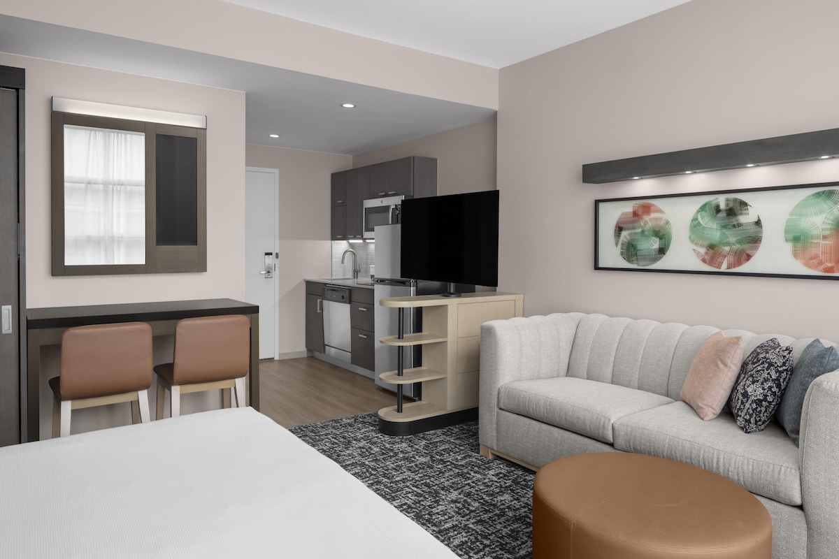 A modern hotel room with a bed, a sofa, a dining area, and a kitchen.