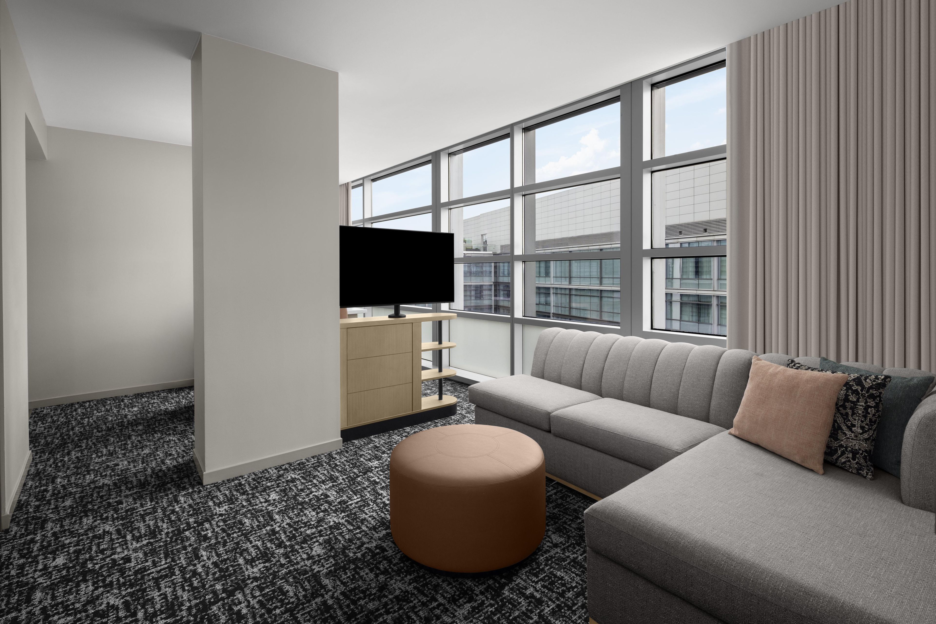 A modern hotel room with a large window, a flat-screen TV, a sofa, a coffee table, and a patterned carpet.