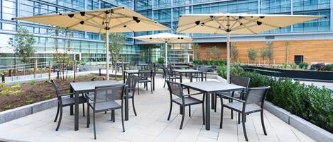 Un área de patio al aire libre con mesas y sillas, grandes sombrillas y un edificio moderno al fondo.