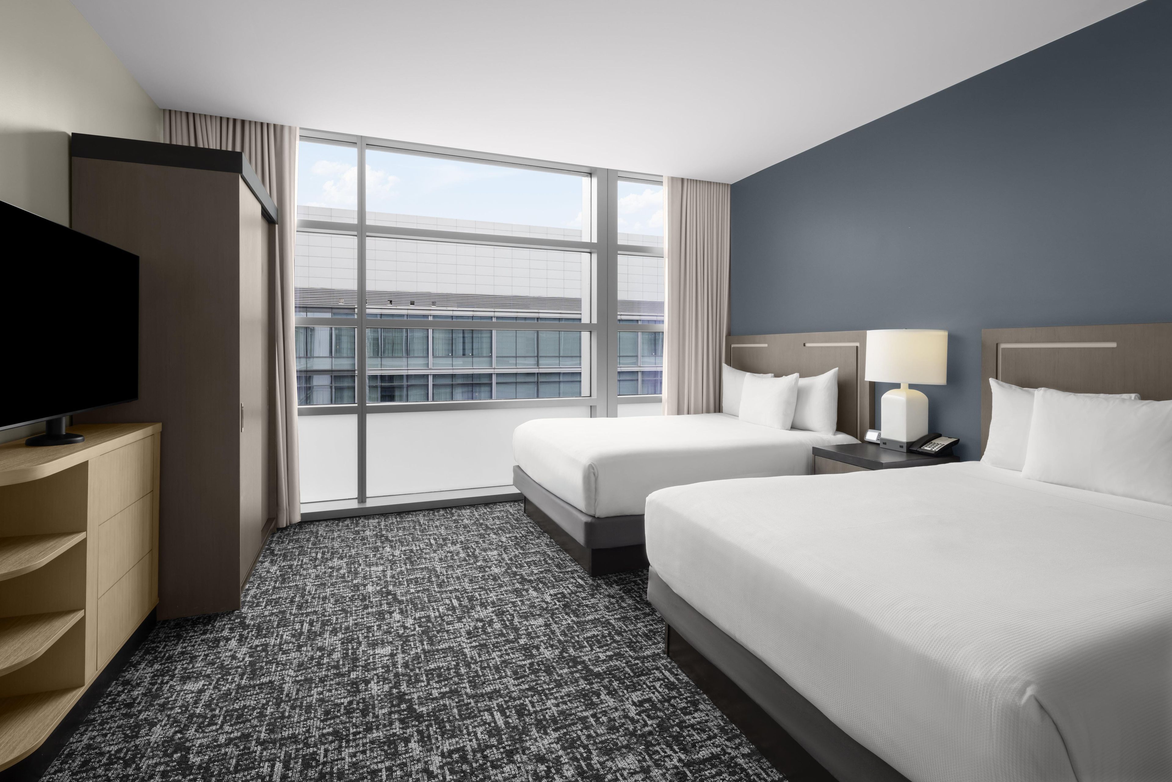 A hotel room with a large window, a flat-screen TV, two beds, a desk, and a telephone.