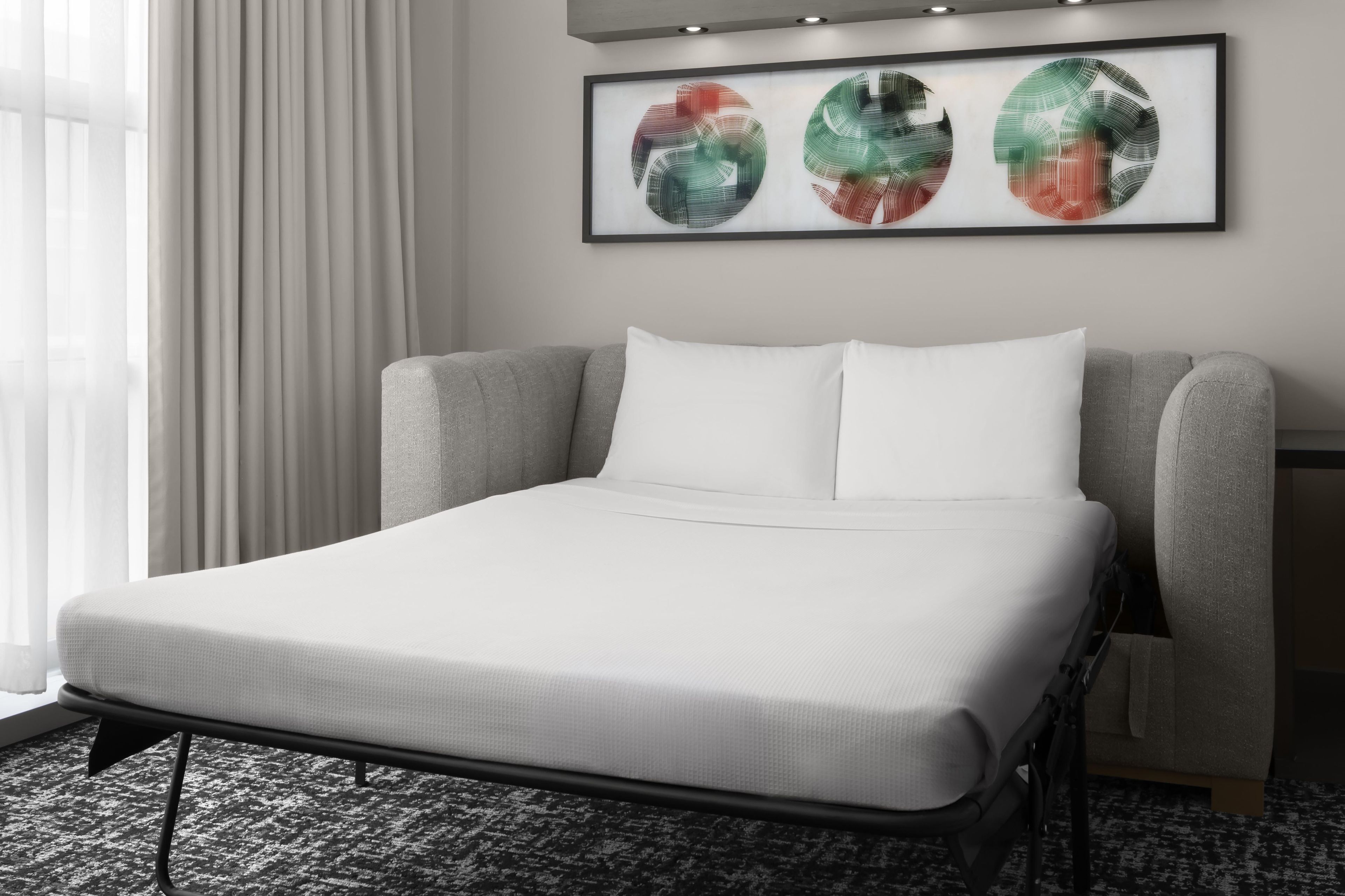 A hotel room with a single bed, a grey upholstered headboard, white bedding, and a framed abstract art piece on the wall.