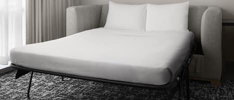 A hotel room with a single bed, a grey upholstered headboard, white bedding, and a framed abstract art piece on the wall.