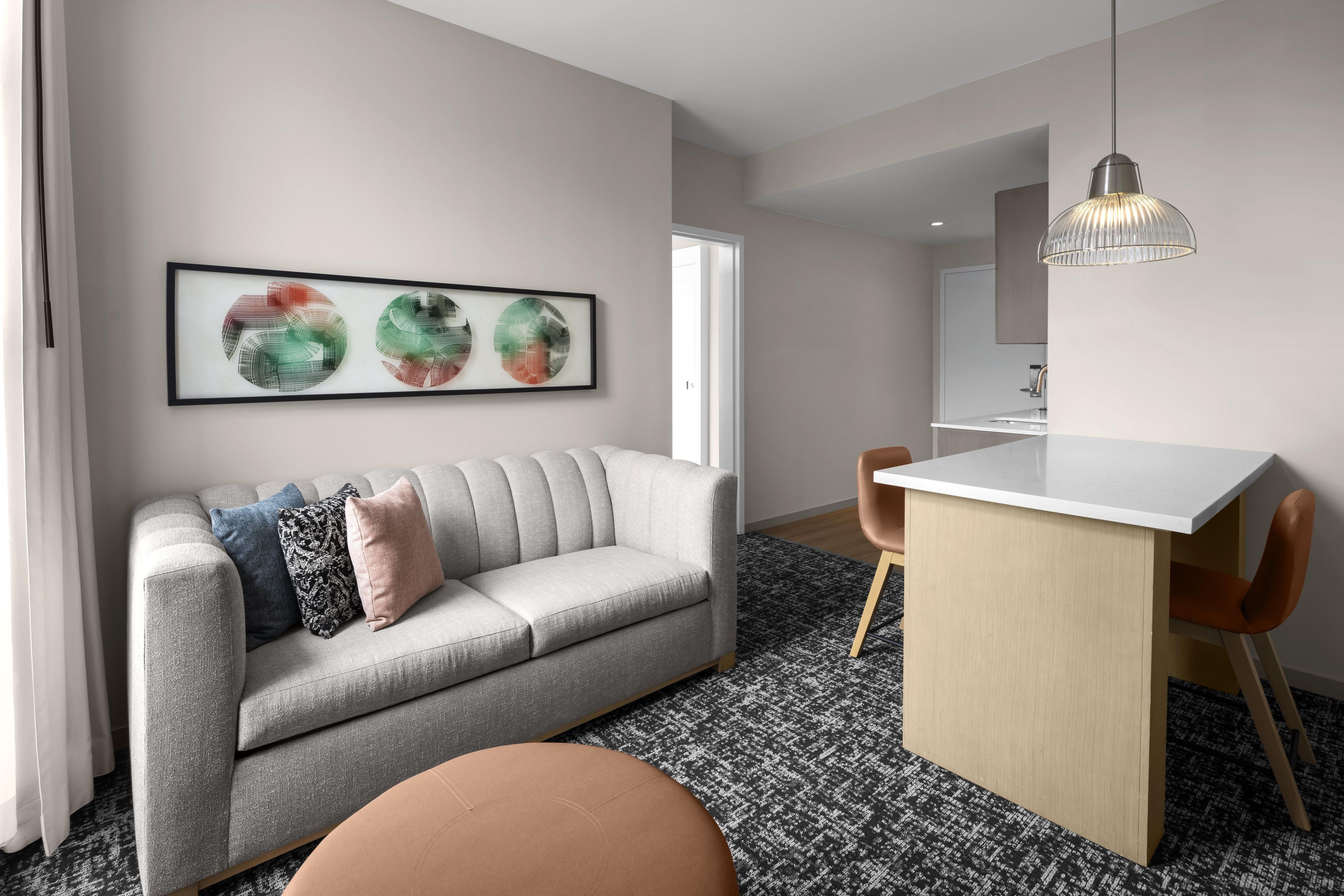 A modern living room with a grey sofa, a round ottoman, a dining area with a white table, and abstract wall art.
