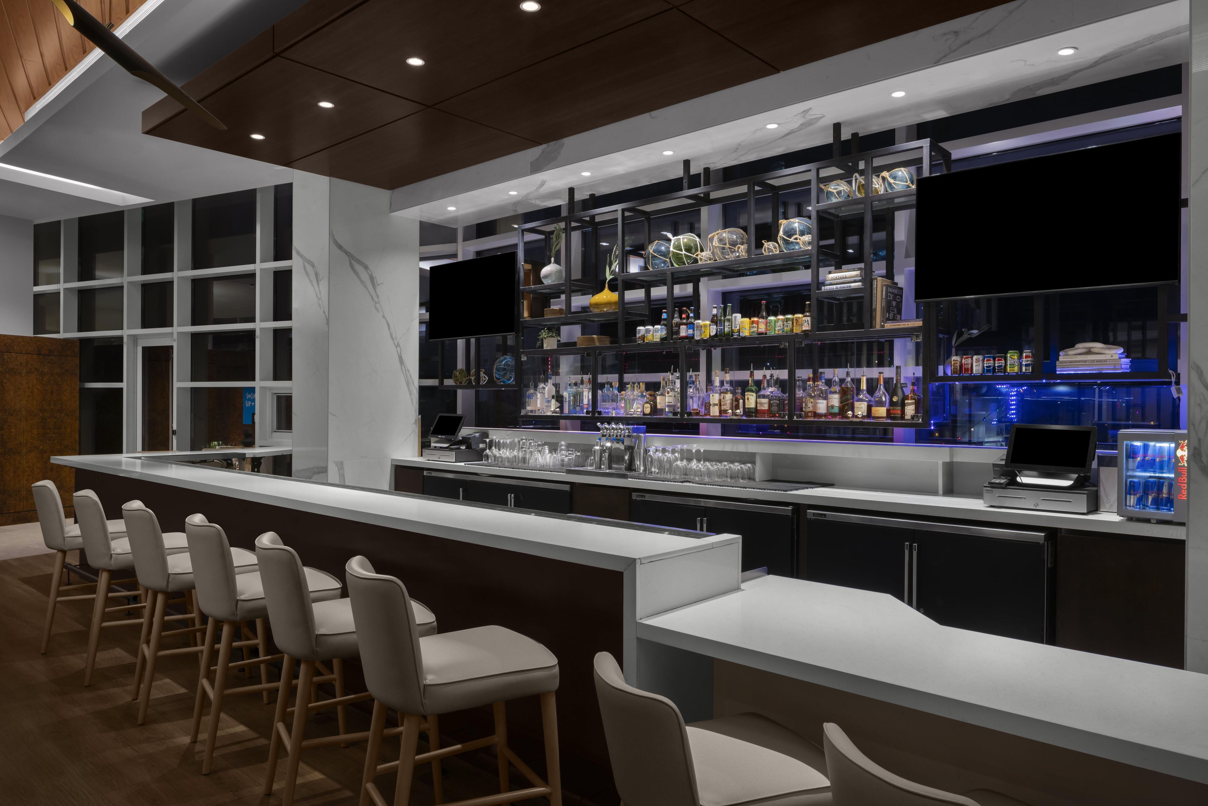 A modern bar area with a long counter, bar stools, and shelves stocked with bottles and glasses.