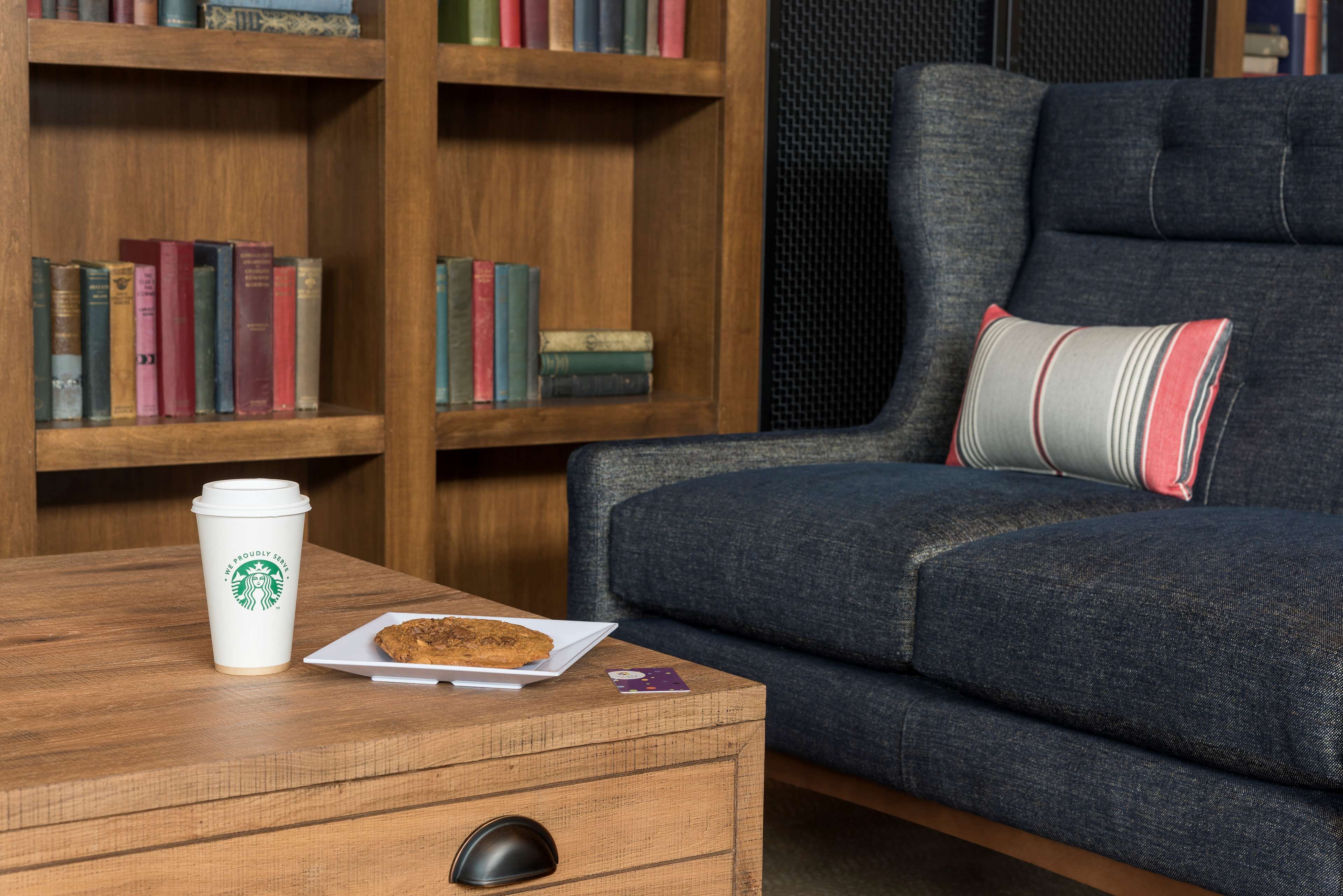 A cozy seating area with a wooden coffee table, a white plate with a cookie, a Starbucks cup, and a smartphone.