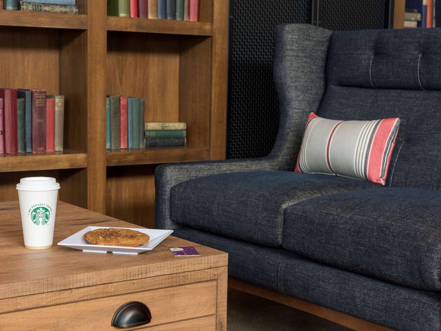 A cozy seating area with a wooden coffee table, a white plate with a cookie, a Starbucks cup, and a smartphone.