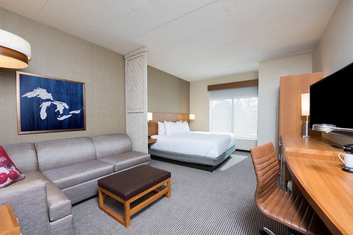 A hotel room with a bed, a sofa, a wooden table, a TV, and a painting on the wall.