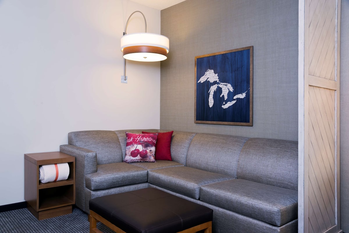 A modern living room with a grey sectional sofa, a wooden side table with a rolled-up white towel, a black ottoman, a wall-mounted light fixture, and a framed abstract art piece.