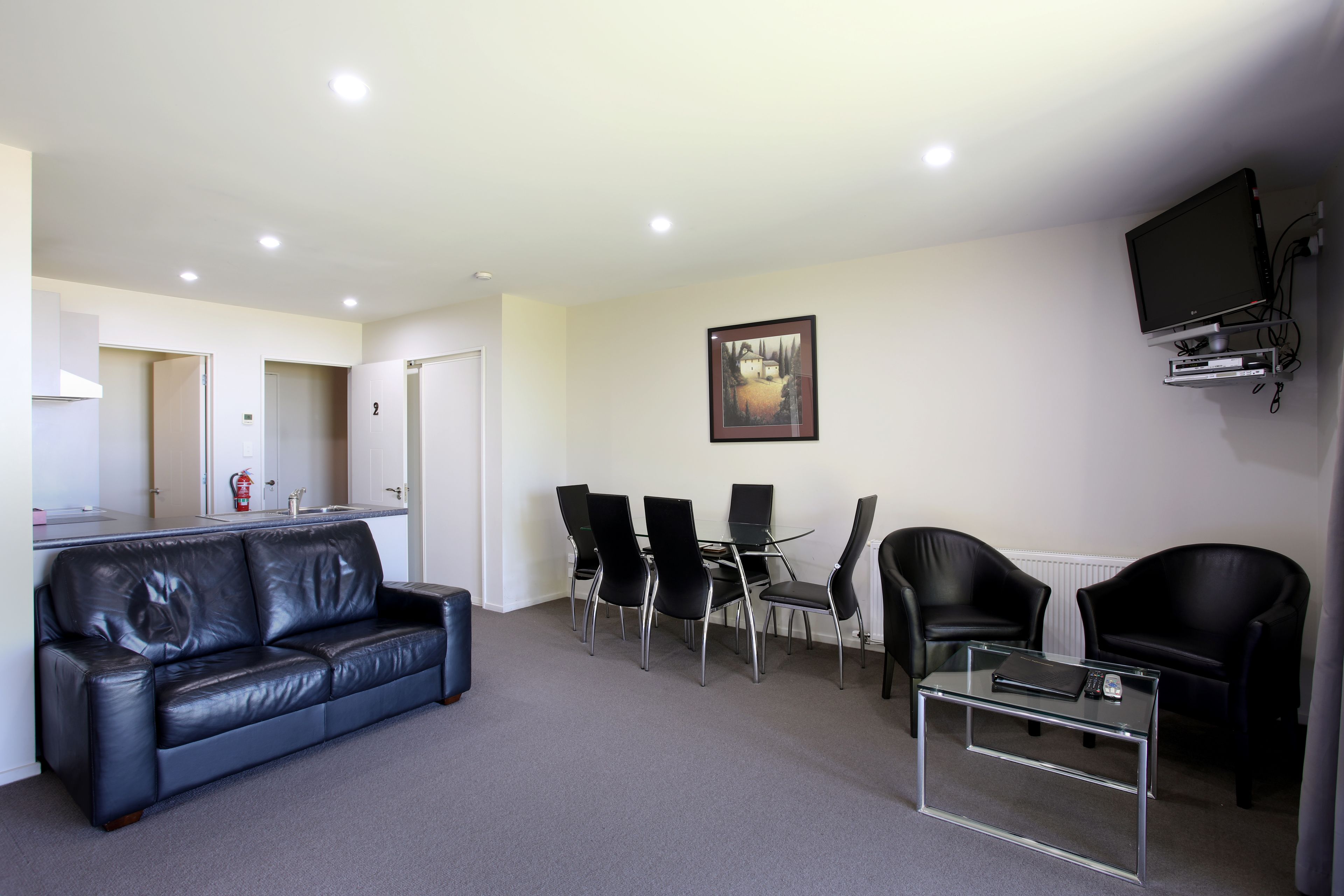 A modern living room with a black leather sofa, a glass coffee table, and a dining area with four chairs.