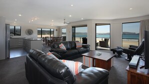 A spacious living area with a black leather sofa, a wooden coffee table, and a television. A kitchen is visible in the background, and large windows offer a view of the beach.