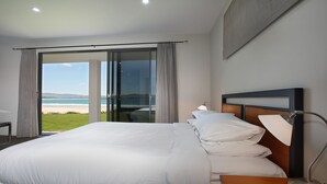 A bedroom with a large bed, a nightstand, a lamp, and a view of the beach through a sliding glass door.