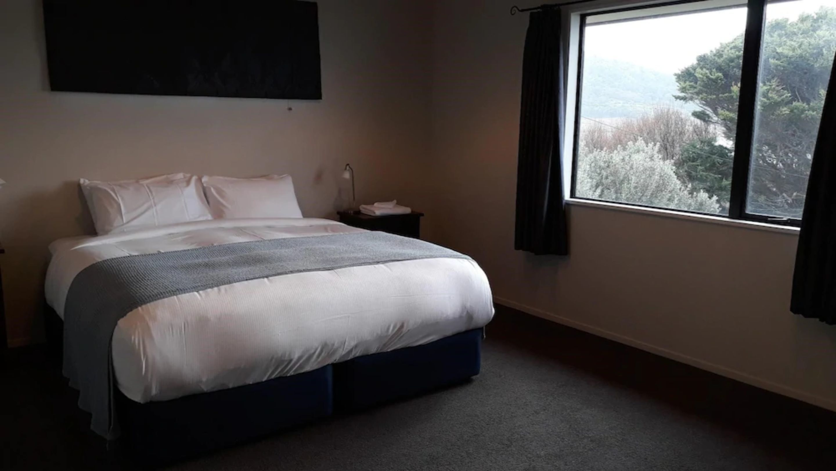 A bedroom with a bed, a nightstand, a window with curtains, and a view of trees.