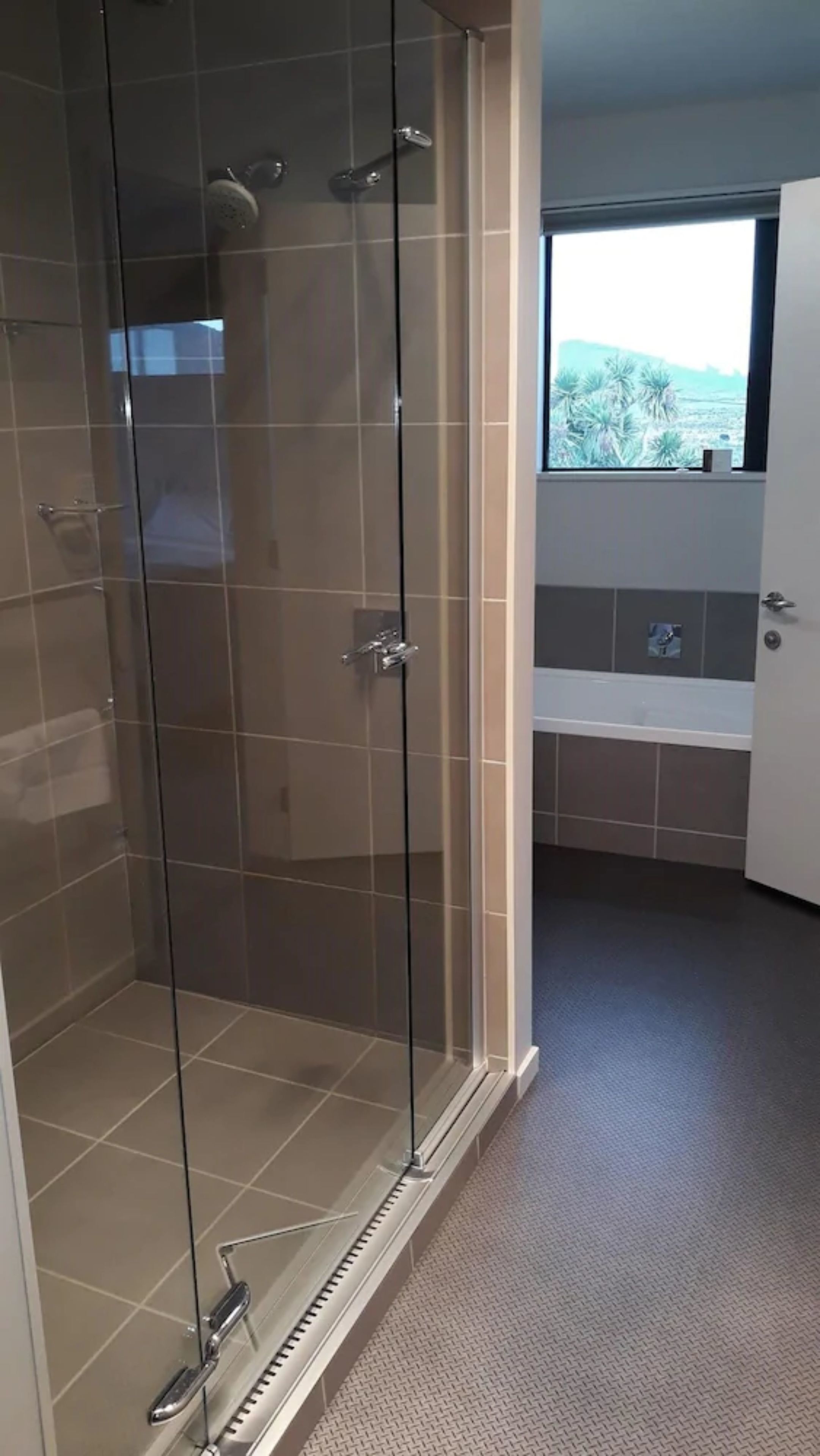 A modern bathroom with a glass-enclosed shower, a bathtub, and a window providing a view of palm trees.