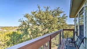 Una terraza de madera con barandas de metal, dos sillas metálicas y vistas a un paisaje arbolado.