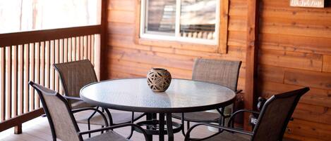 Una terraza de madera con una mesa redonda y cuatro sillas, una ventana y un muro de madera con un letrero.