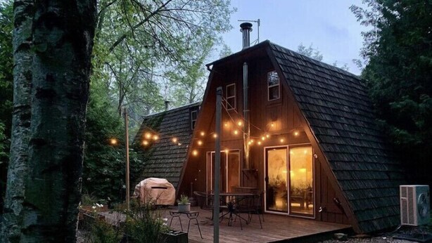 Una cabaña de madera con techo inclinado, un porche con mobiliario exterior y luces colgantes.
