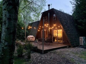 Une cabane en bois avec un toit en pente, une terrasse dotée de meubles d’extérieur et illuminée de guirlandes lumineuses.