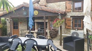 Een terras buiten met een stenen gebouw, een bord voor de barbecueplaats, zitplaatsen buiten en een airconditioning.