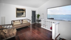 Een ruime woonkamer met een houten vloer, een bank, een bureau met een laptop en uitzicht op zee door een groot raam.