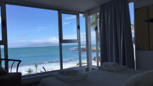 Een hotelkamer met een groot raam dat uitzicht biedt op de oceaan en het strand. Er staat een bed met witte lakens en een stoel.