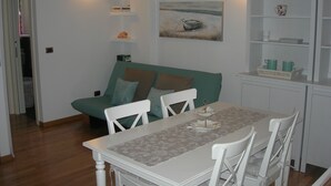 Ein Essbereich mit einem weißen Tisch, Stühlen und einem grünen Sofa. An der Wand hängt ein Gemälde und es gibt Regale mit Gegenständen.