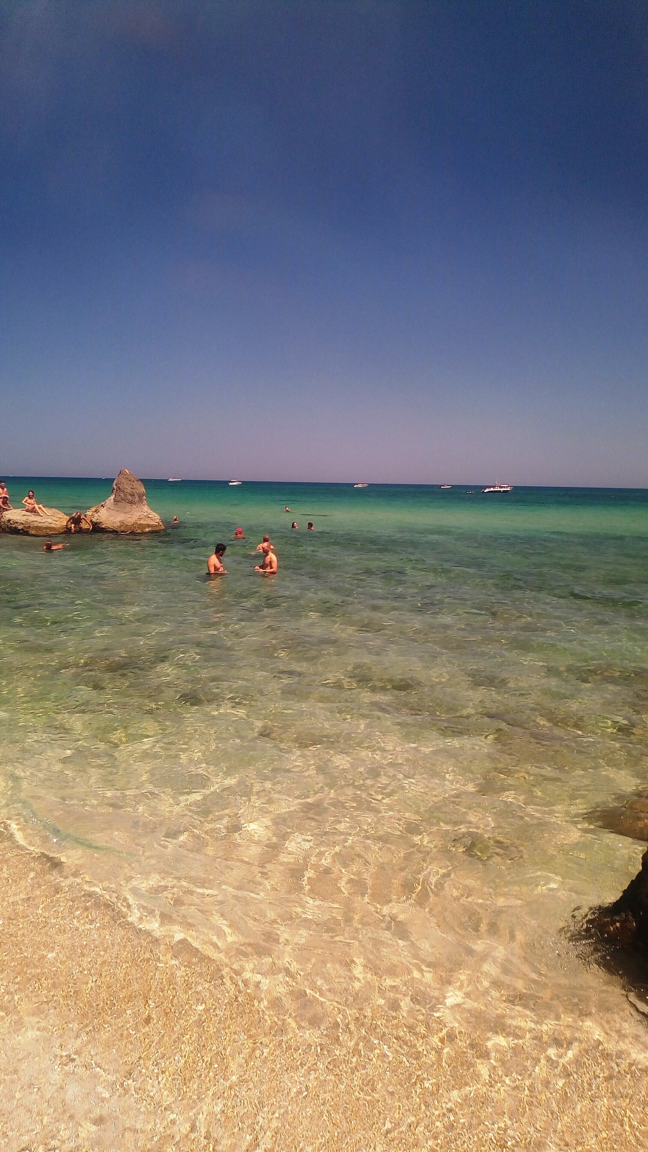 Una playa con aguas cristalinas y poco profundas, algunas personas nadando y un promontorio rocoso a lo lejos.