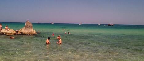 Una playa con aguas cristalinas y poco profundas, algunas personas nadando y un promontorio rocoso a lo lejos.