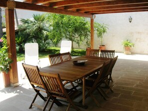 A wooden table with six chairs under a wooden pergola, surrounded by potted plants and a white wall.